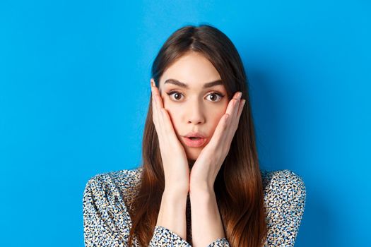 Close-up portrait of surprised cute woman holding hands on cheeks and gasping, stare at camera shocked, standing against blue background.