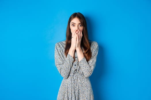 Shocked european girl gasping and covering mouth with hands, witness something and look startled, standing worried on blue background.