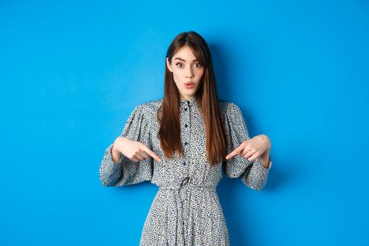 Excited beautiful woman in dress say wow, pointing fingers down and look amused, checking out promotion, standing on blue background.