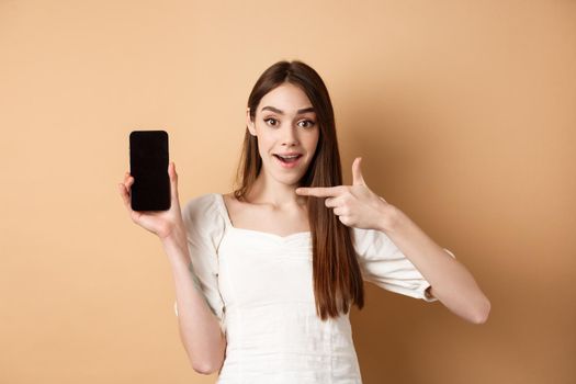 Cheerful woman pointing finger at empty phone screen, looking excited, standing on beige background.