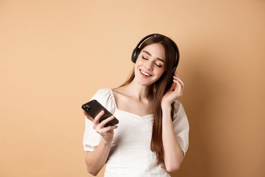 Happy girl listening music in headphones and smiling, holding smartphone in hand, beige background.