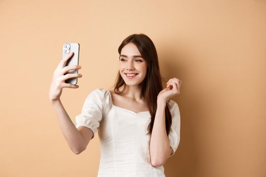 Beautiful woman taking selfie on smartphone, posing for phone photo, standing on beige background.