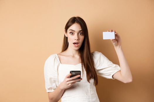 Excited woman showing plastic credit card and using mobile phone app, drop jaw and gasping amazed, checking out bank offer, beige background.