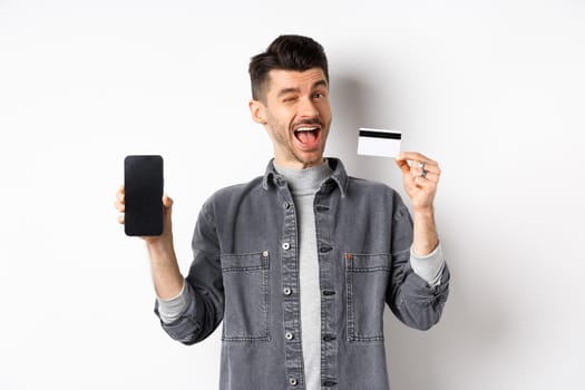 Cheerful handsome guy showing good deal, empty smartphone screen and plastic credit card, winking at you and smiling, recommending offer, white background.