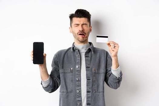 Sad crying man show empty mobile phone screen and plastic credit card, standing miserable on white background.