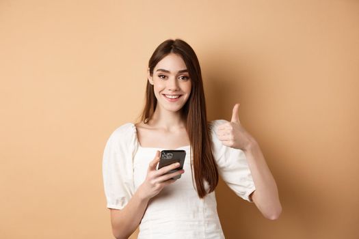 Online shopping concept. Smiling happy woman showing thumb up after using smartphone, holding phone and say yes, standing on beige background.