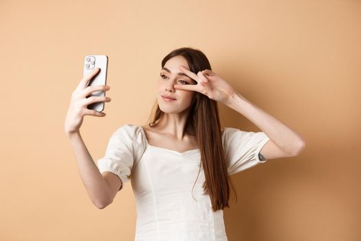 Cute woman taking selfie on smartphone, showing v-sign near eye, posing for social media photo, standing on beige background.