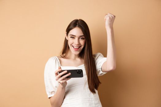 Girl winning on mobile phone. Happy woman raising hand up and scream yes with joy, achieve goal in smartphone app, standing on beige background.