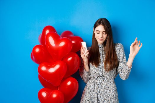 Portrait of attractive young woman holding smartphone and dancing near Valentines red balloons, standing on blue background.