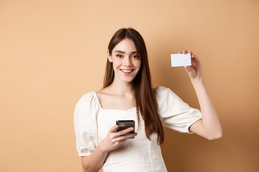 E-commerce concept. Smiling woman showing plastic credit card while shopping online on mobile phone, standing against beige background.