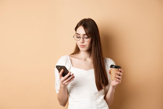 Busy girl in glasses reading message on mobile phone and drinking coffee takeout, standing on beige background.