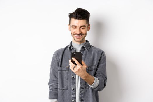 Handsome man reading message on phone and smiling. Guy holding smartphone and looking at screen, standing on white background.