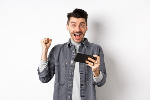 Excited man making money on mobile phone and rejoicing, raising hand up and scream from happiness and joy, standing with smartphone on white background.