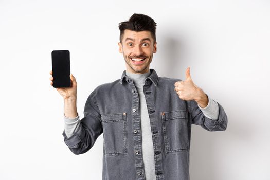 Cheerful stylish man showing empty smartphone screen and thumbs-up, concept of online shopping and e-commerce.