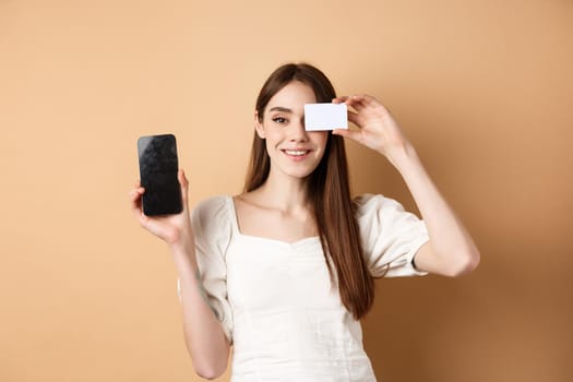 Cheerful young woman showing plastic credit card and empty mobile phone screen, smiling pleased at camera, standing on beige background.