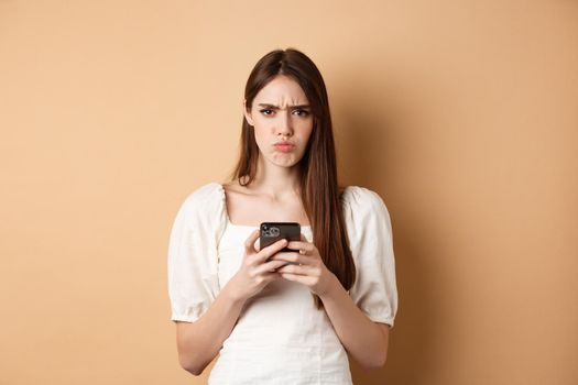 Disappointed girl with smartphone frowning, pucker lips upset, reading bad news on phone, standing on beige background.