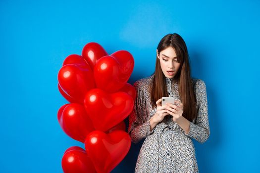 Valentines day. Portrait of young woman standing near red romantic balloons, looking surprised at smartphone screen, blue background.