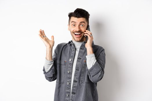 Excited man talking on cellphone and hear good news, rejoicing and raising hand with happy smile, standing against white background.