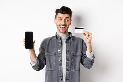 Excited handsome man showing plastic credit card and empty smartphone screen, advertising application, standing on white background.