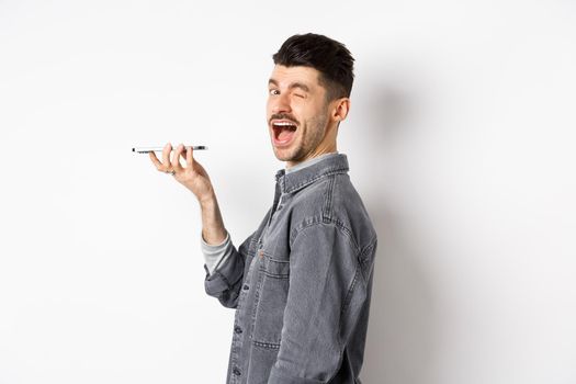 Happy guy winking at looking at camera while using voice translator app or talking on speakerphone, holding phone close to mouth, standing on white background.