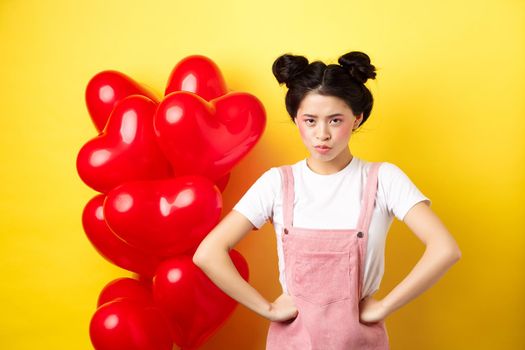 Valentines day and relationship concept. Disappointed asian girlfriend sulking, looking offended at camera, mad at boyfriend, standing near heart balloons, yellow background.