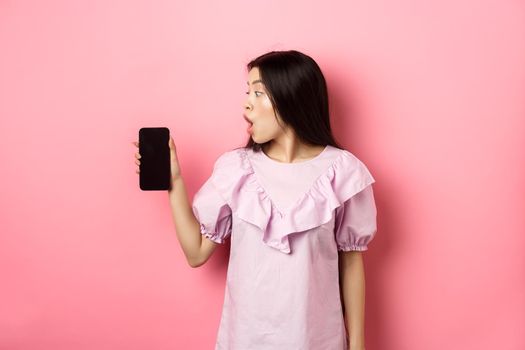 Wow look here. Excited asian girl look at smartphone screen with amazed face, checking out online offer, standing against pink background.