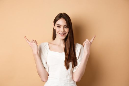 Beautiful young woman in dress pointing fingers sideways, show two ways, giving choice and smiling, standing on beige background.