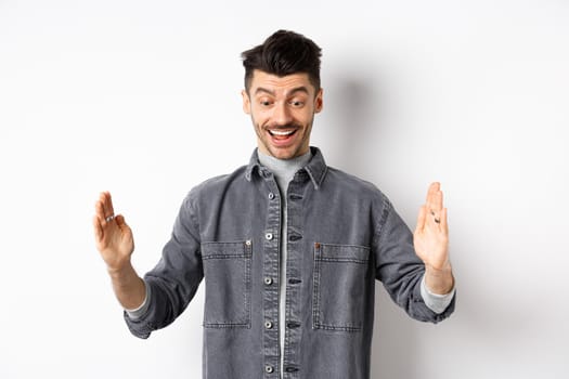 Excited caucasian man showing big size object, shaping large thing and smiling amazed, standing on white background.