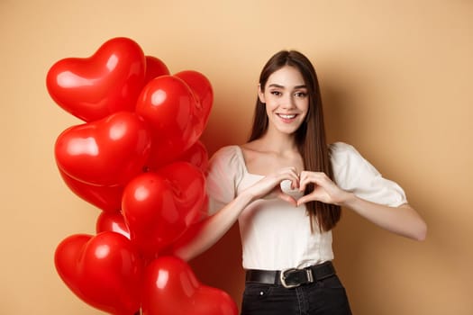 Lovely caucasian girl express love and tenderness, showing heart gesture and smiling, standing near Valentines day balloons, beige background.