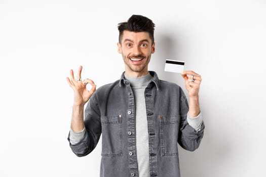 Very good. Smiling guy with plastic credit card showing okay sign, smiling satisfied, recommend bank, standing on white background.