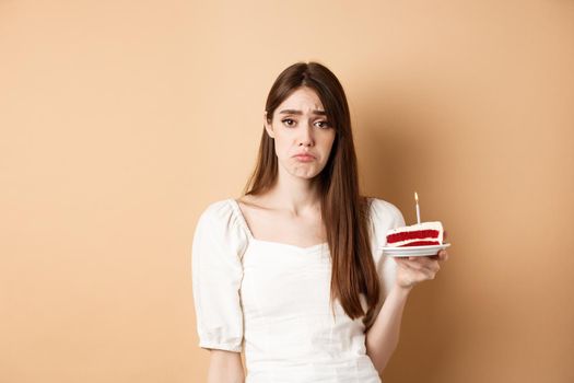 Sad birthday girl holding cake with candle and look upset, feeling lonely on her bday, standing alone on beige background.