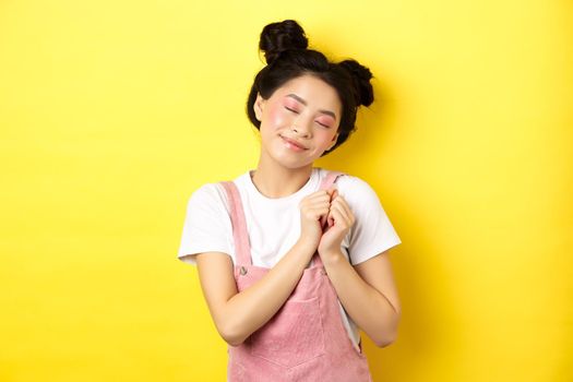Cute asian girl with makeup, close eyes and remember beautiful moment, holding hands on heart daydreaming, standing on yellow background.
