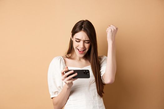 Euphoric happy winner playing mobile video game, shouting yes with joy and making fist pump, winning online, looking at smartphone screen, beige background.