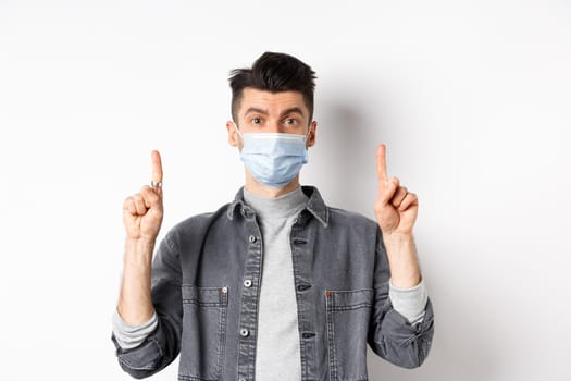 Pandemic lifestyle, healthcare and medicine concept. Stylish modern man in medical mask showing advertisement, pointing fingers up at logo, standing on white background.