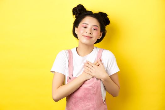Beautiful teen asian girl in love, holding hands on heart and smiling touched, looking with affection, standing on yellow background.