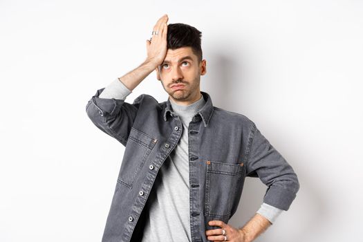 Annoyed young man looking tired or bothered, roll eyes and slap forehead, making facepalm from something stupid, standing on white background.