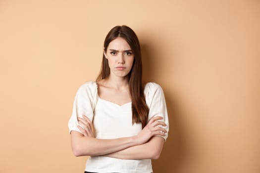 Angry girlfriend cross arms on chest and frowning, looking offended or disappointed, sulking at you, standing mad on beige background.