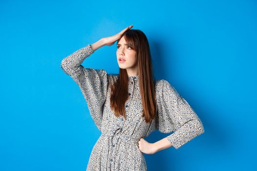 Troubled young woman in dress slap forehead and roll eyes up, standing tired and bothered with work, blue background.