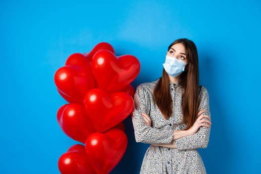 Covid-19, quarantine and health conept. Dreamy beautiful girl in face mask and dress, looking at upper left corner pensive, standing near Valentines day balloons, blue background.