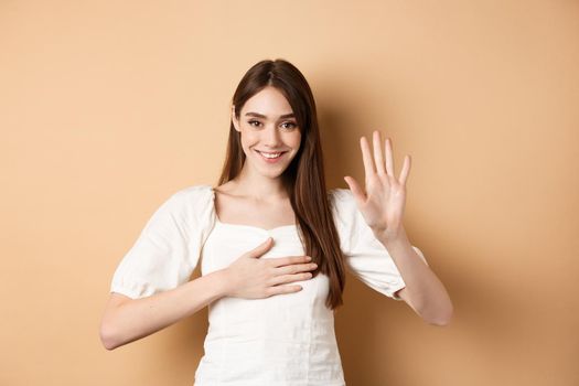 Cute smiling girl make promise, put hand on heart and tell truth, being honest, swearing to you, standing on beige background.