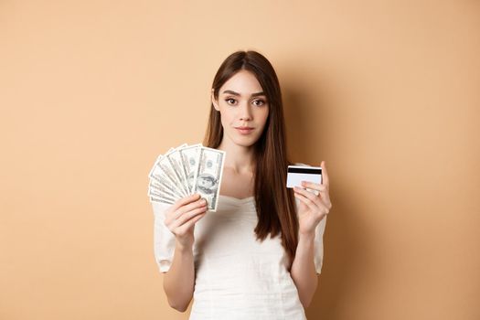 Pretty young woman in white blouse showing dollar bills and plastic credit card, contactless payment vs cash, standing on beige background.