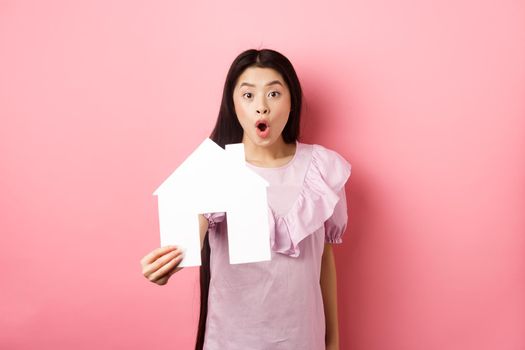 Real estate and insurance concept. Excited asian woman showing paper house cutout and look amazed, special offer, standing on pink background.