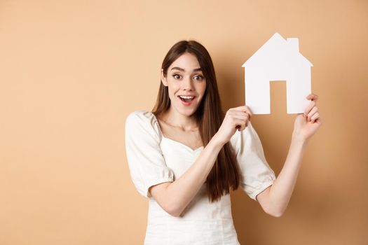 Real estate. Excited woman got new apartment, showing paper house cutout and smiling happy, standing on beige background.