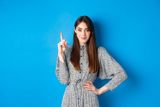 Young beautiful lady in dress with natural long hair, showing number one with finger and smiling, standing against blue background.