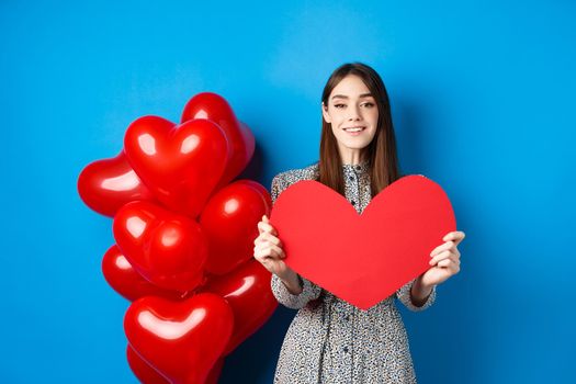 Valentines day. Lovely young woman in dress celebrating lovers holiday, showing valentine card and smiling, standing near red heart balloons on blue background.