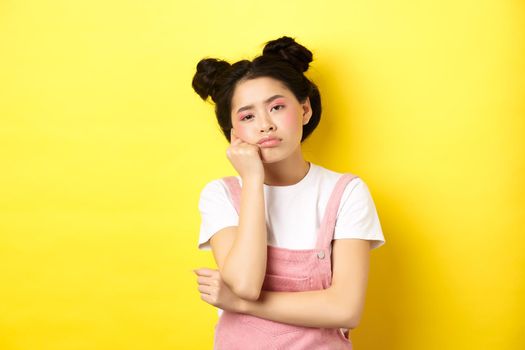 Sad and bored teen asian girl standing alone, looking at camera indifferent, staring with boredom, yellow background.