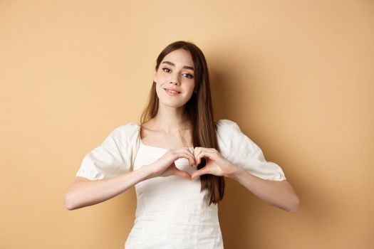 Beautiful caucasian woman say I love you, showing heart gesture and smile at camera, beige background. Concept of romance and Valentines day.