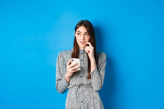 Pensive cute woman thinking how to answer on message, looking aside thoughtful and holding smartphone, standing in dress on blue background.