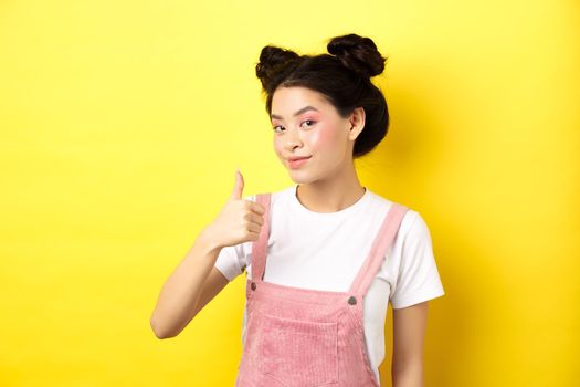 Beautiful asian girl with bright pink makeup, showing thumb up in approval and smiling satisfied, recommending promo, yellow background.