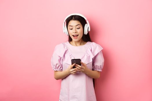 Happy beautiful woman in wireless headphones watching video on smartphone, looking at phone amused, standing against pink background.